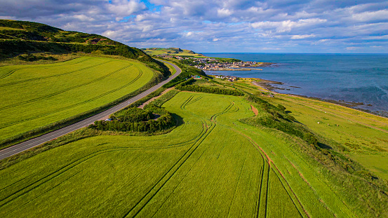 AERIAL: Scenic view of big wheat field and lovely village on coast of North Sea. Road winding along picturesque Scottish coastline in rich shades of green and blue with beautifully landscaped farmland