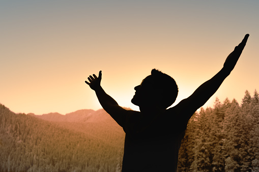 Rear view of a woman's silhouette on top of a hill as she looks at the sun and valley with arms raised.