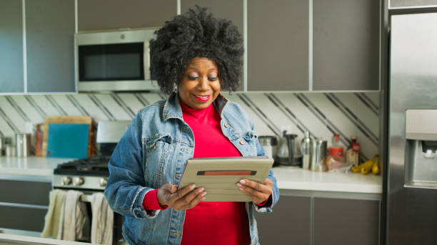 istock Mature African American Woman Smiling at Digital Tablet while Standing inside Kitchen 2120866034
