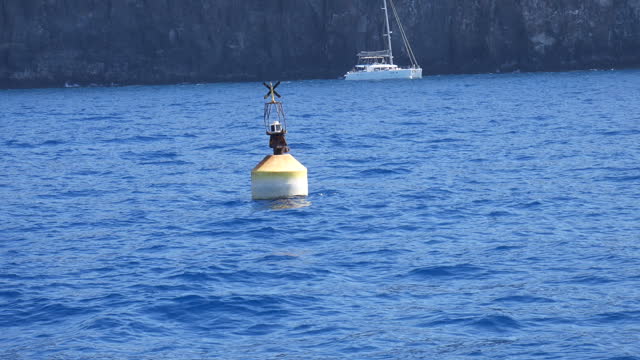Navigation buoy floating on blue ocean water, sailboat and rocky coast in the background. Marine safety, coastal navigation, sea marker.