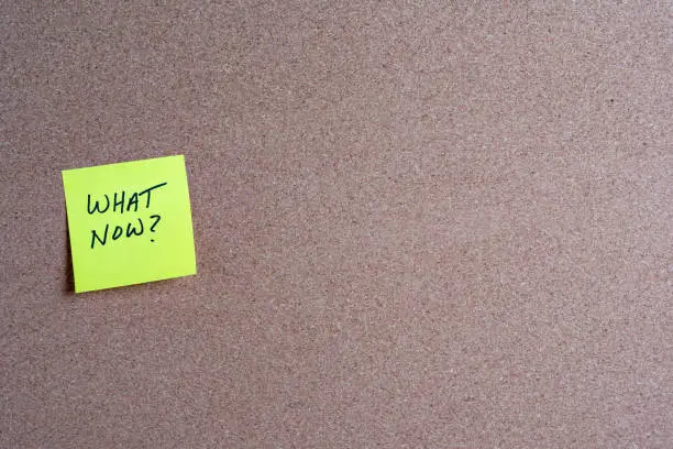 The words What Now on a yellow sticky note posted on a corkboard