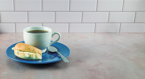 Coffee in white porcelain cup on blue plate with bagel and cream cheese on pink tinted counter top with white subway tile back splash