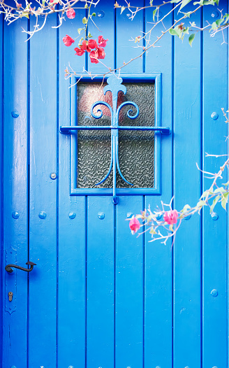 Blue door featuring a window with glass, with bouganvillea branches and petals. Typical door of the fishing villages of the Mediterranean coast, Catalonia, Spain.