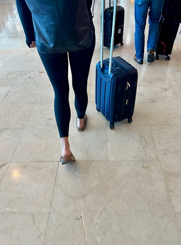 Rear view of a woman's back and legs wearing a backpack, black leggings and slip-on shoes, wheeling a small, hard-shell, carry-on suitcase past other travelers in the Cancun airport