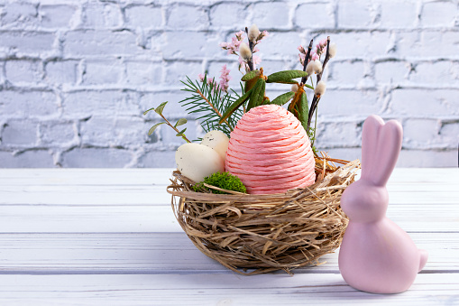 Easter decoration - nest with big pink egg made of rope and little speckled eggs, branches and flowers, pink figurine of bunny on the white wooden table at the brick wall.