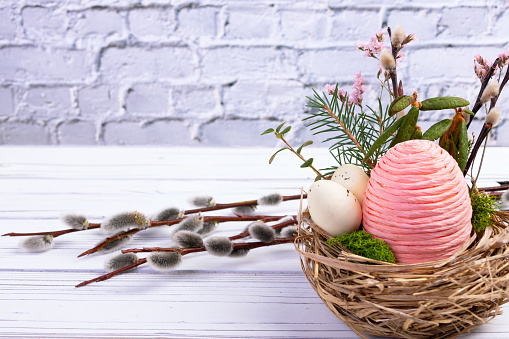 Easter decoration nest with big pink egg made of rope and little speckled eggs, branches and flowers, pussy willow behind it, on the white wooden table at the brick wall.