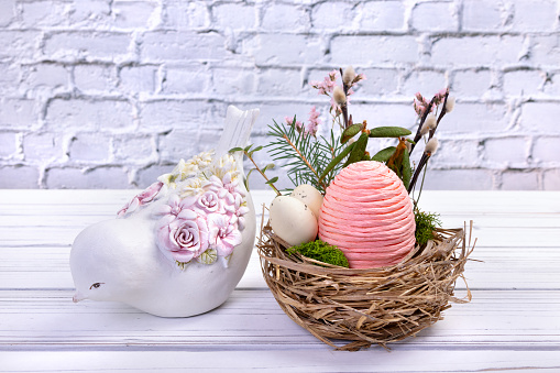 Easter decoration nest with big pink egg made of rope and little speckled eggs, branches and flowers, figurine of clay painted bird on the white wooden table at the brick wall.