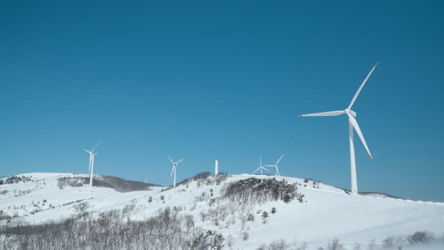 Industrial Windmills Farm or Wind Turbines Rotate Blades Atop Snow-covered Winter Mountains Against Clear Sky in Alps