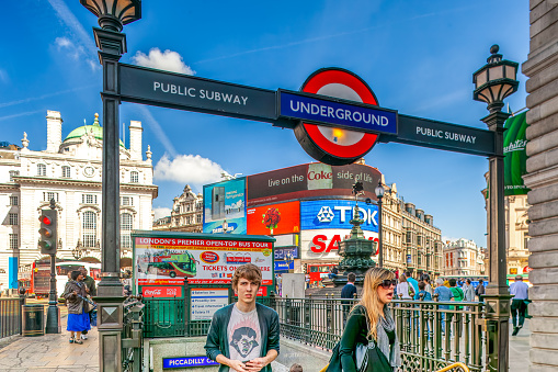 Pedestrians walking past the famous Piccadilly Circus station on a sunny day.