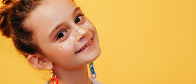 Kid smile. Joyful child. Cheerful happy little girl with fake sparkling freckles in colorful handmade earrings good mood isolated on yellow copy space background.
