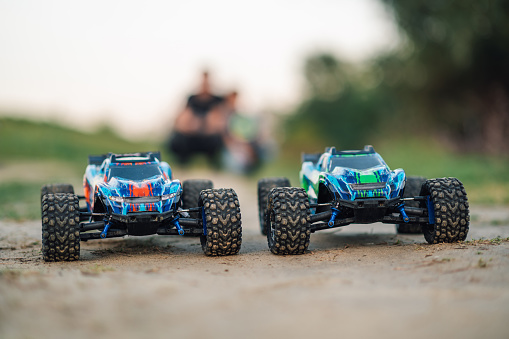 Selective focus on two electric toy electric, standing on a dirt road. They are ready for a wild race. In the blurred background, there are two figures of a father and his son.