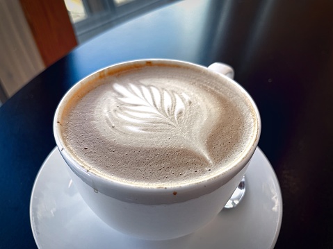 Cup of cappuccino with artful foam design on a saucer, placed on a dark wooden table with soft natural light.