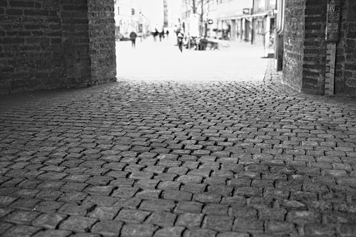 Cobblestone street view in monochrome, leading to an urban scene with blurred pedestrians in the background.