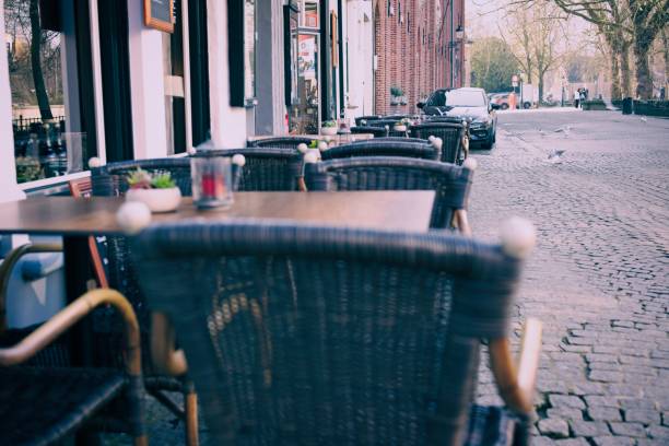 istock Empty street cafe with wicker chairs on a cobblestone sidewalk, urban European setting with parked cars and trees. 2121197532