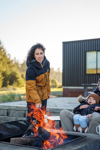 A woman of Eurasian descent stands next to a fire pit, roasting a marshmallow while enjoying a cold day outside with family at a rental house at the Oregon coast.