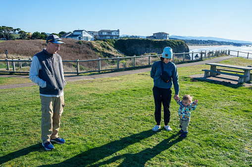 An active senior man talks with his adult daughter and her toddler son while enjoying time outside exploring the Face Rock State Scenic Viewpoint in Bandon, Oregon while on a family road trip in the winter.