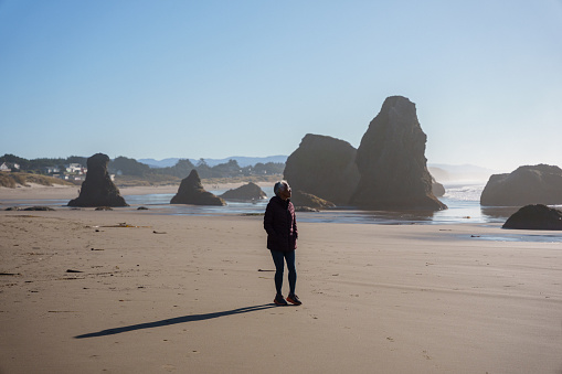 An active senior woman of Hawaiian and Chinese descent who is wearing a winter jacket, takes a relaxing walk along an empty beach in Bandon, Oregon with beautiful rock formations while on a winter trip.