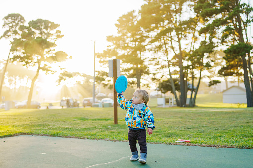 A cute one year old Eurasian boy holds up a frisbee while playing disc golf with his family at a park in Oregon on a sunny and cool afternoon.
