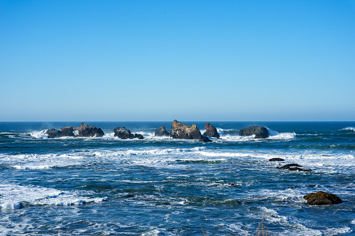 View of ocean waves crashing onto large rock formations at the Face Rock State Scenic Viewpoint in Bandon, Oregon on a sunny day with no people.