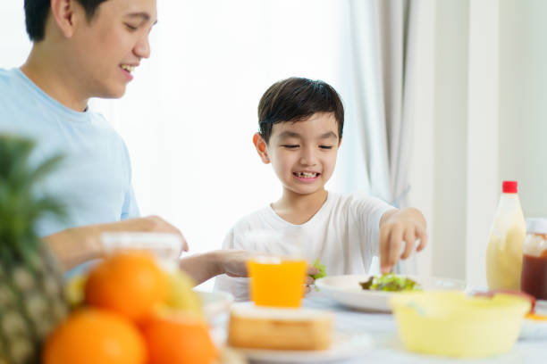 Lovely happy Asian family, father and son, having a healthy breakfast together with a breads, salad, fruits, and fried egg. Father teach his little son making an easy and healthy breakfast.