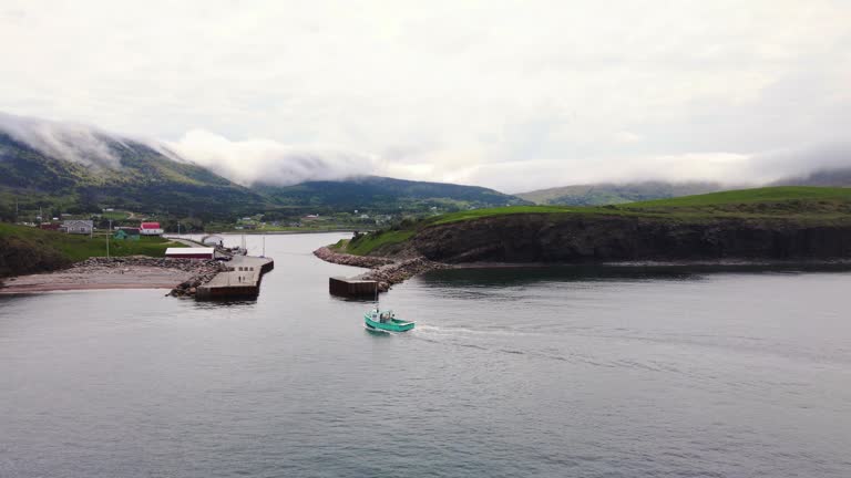 Drone shot of a small commercial lobster fishing boat coming back into the harbour after a long day of working revealing the beautiful landscape of the Cape Breton Coast in Nova Scotia, Canada.