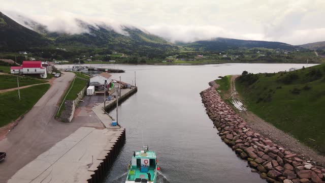 Drone shot following a small commercial lobster fishing boat coming in after a long day revealing a beautiful small village with mountains in the background on Cape Breton Island, Nova Scotia.
