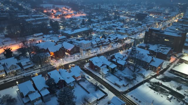 American town during snow flurries at dawn. High aerial above quaint main street lined with Christmas lights and fresh snow.