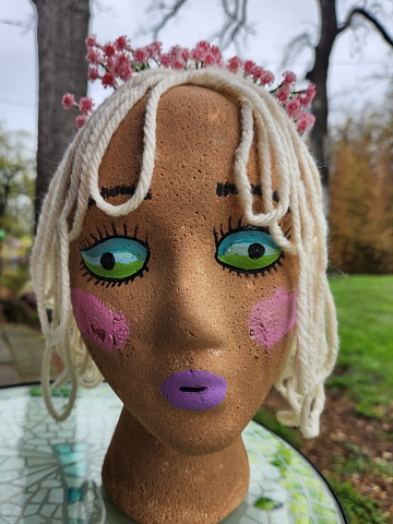 A craft styrofoam head painted with dark skin and blue and green reptile like eyes. The head has pink and purple makeup and has beige yarn for hair. There is a flower crown in the hair and creepy trees in the background.