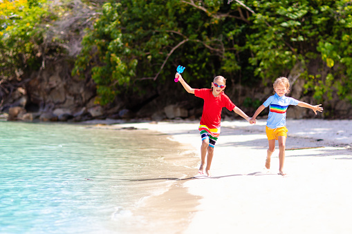 Kids run and play on tropical beach. Children swim and jump in sea on summer family vacation. Sand and water fun, sun protection for young child. Little boy running and jumping at ocean shore.