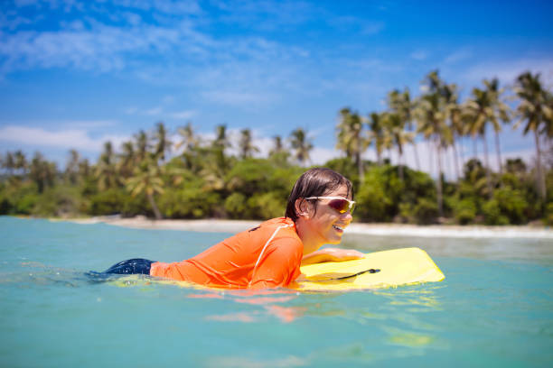 Teenager boy surfing on tropical beach in Asia. Child on surf board on ocean wave. Active water sports for kids. Kid swimming with body board. Young surfer in exotic sea. Swim and eye wear for kids.
