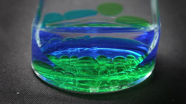 Macro view of a glass beaker as green and blue bubbles are added, abstract science art.