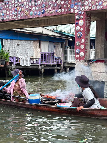 Bangkok, Thailand - February, 15 2024: Stock photo showing  view of river boat touring the canals and waterways of Khlong Damnoen Saduak a canal link to Tha Chin and Mae Klong rivers. Locals barbequing meat on metal drum grill aboard a flat bottomed, wooden boat.