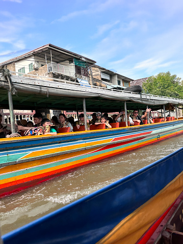 Bangkok, Thailand - February, 15 2024: Stock photo showing  view of river cruise tour boat with tourists wearing safety jackets, touring the canals and waterways of Khlong Damnoen Saduak a canal link to Tha Chin and Mae Klong rivers.