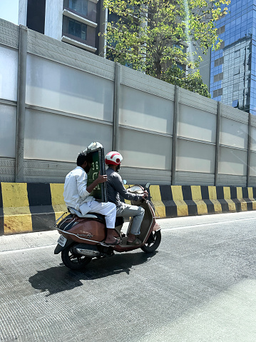 Mumbai, Maharashtra, India - March, 15 2024: Stock photo showing two people riding motorcycle with the pillion passenger holding large cumbersome television screen on Indian main road with black and yellow striped concrete safety barrier.