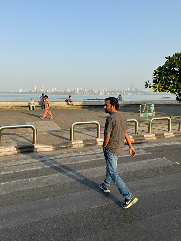 Marine Drive, Mumbai, Maharashtra, India - March, 16 2024: Stock photo showing close-up view of zebra crossing with painted road markings on tarmac surface being crossed by Indian man wearing casual clothing.
Crossing multilane highway running along coastline at Marine Drive, Mumbai, India with a manmade, sea defence of a concrete, seawall, which has been constructed to prevent flooding of the promenade of  during high tide.