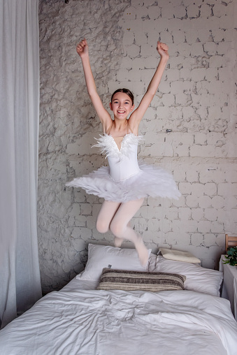 Joyful Young Ballerina Jumping on Bed. Young girl in white swan tutu leaps joyfully on bed, fooling around, smile radiating pure happiness in cozy bedroom setting.