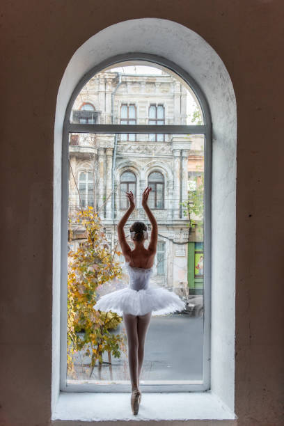 Ballerina poses en pointe, white tutu within an arched window frame, juxtaposing the refined beauty