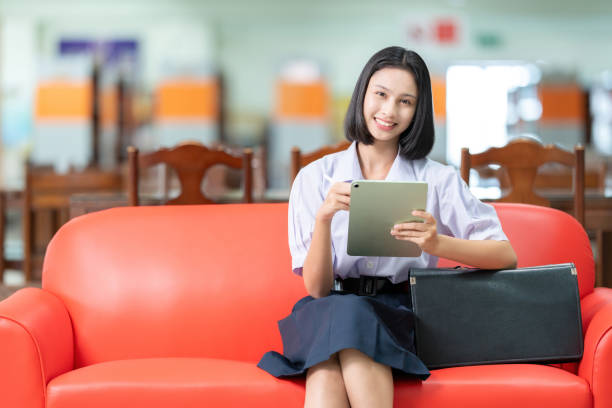 Asian female student with short hair smiles cutely Sitting on the red sofa in the library Use a laptop to research. Next to it is a school bag. Wear a uniform in the library to gain knowledge. Asian female student with short hair smiles cutely Sitting on the red sofa in the library Use a laptop to research. Next to it is a school bag. Wear a uniform in the library to gain knowledge. cutely stock pictures, royalty-free photos & images