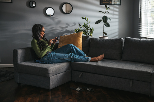 Young woman resting on the couch in the living room texting and scrolling mobile phone social media.