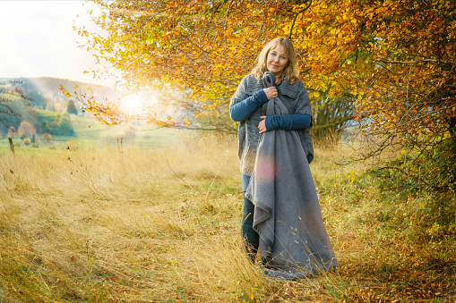 A woman of 40 years in autumn nature landscape. The blonde woman wears clothes made of wool. She has a blanket.
