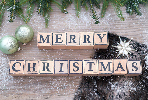 Wooden cubes with letters form the English text Merry Christmas. They are lying on a wood background. Fir branches and Christmas balls are next to it.