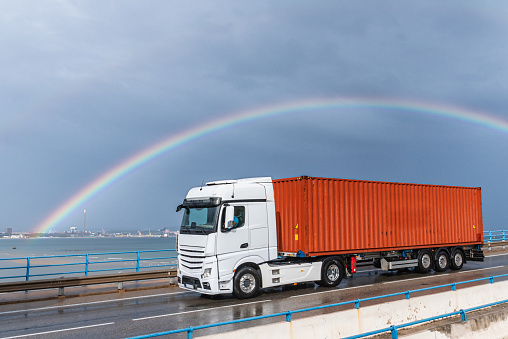 Truck with maritime container driving on a wet road and under a rainbow.