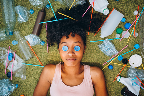 A woman lies amidst a vivid assortment of discarded plastics, using bottle caps as eyes to symbolize the urgent gaze on environmental issues. The striking image serves as a creative call to action against pollution.