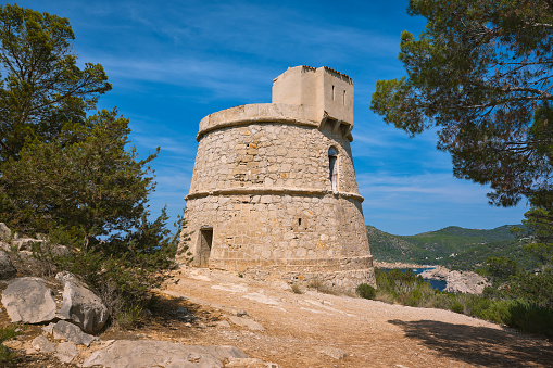 Wide-angle view of Torre des Molar, also known as Torre de Balansat, an 18th-century watchtower located on the northern coast of Ibiza, not far from Port de Sant Miquel (Sant Miquel de Balansat). The dazzling warm light of a Mediterranean summer afternoon, picturesque clouds, lush pine trees, rosemary bushes and juniper hedges. The cove and verdant hills of Benirràs can be seen on the background. High level of detail, natural rendition, realistic feel. Developed from RAW.