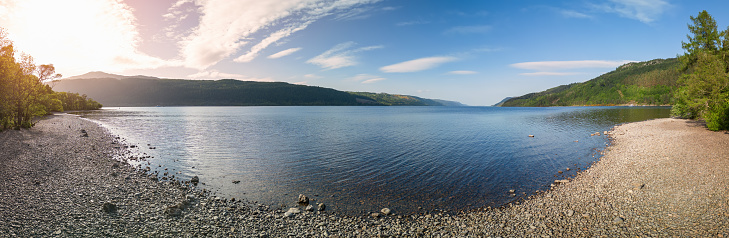 Drone panorama showcases Loch Ness in its full glory, with the sunlight casting a soft glow over the serene lake, pebbled beaches, and the surrounding verdant hills, Scottish Highlands