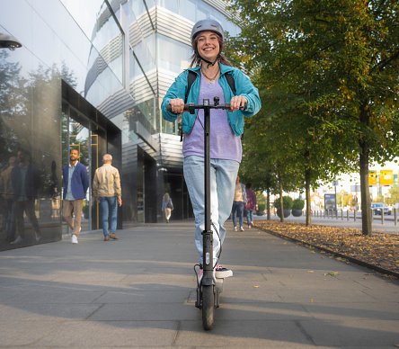 Young Caucasian woman wearing a helmet and smiling while riding her electric scooter in the city.