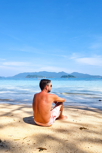 A man in casual clothing sits on the sandy beach, his gaze fixed out to the vast expanse of the ocean before him. Koh Wai Thailand