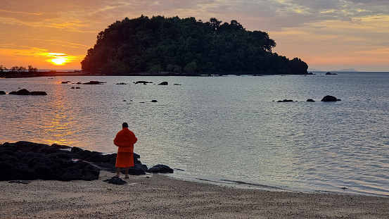 A Thai monk figure stands on a sandy beach as the sun sets, casting a warm glow over the horizon. Koh Libong Thailand