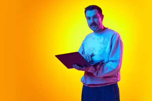 istock Man in sweatshirt standing with laptop on gradient yellow orange background in neon light. Online communication, remote work 2122113838
