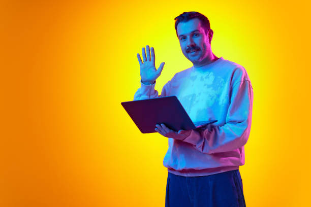 istock Man in tie-dye sweatshirt waves at laptop screen, having online video call on gradient yellow orange background in neon light. Online meeting with colleagues 2122113849